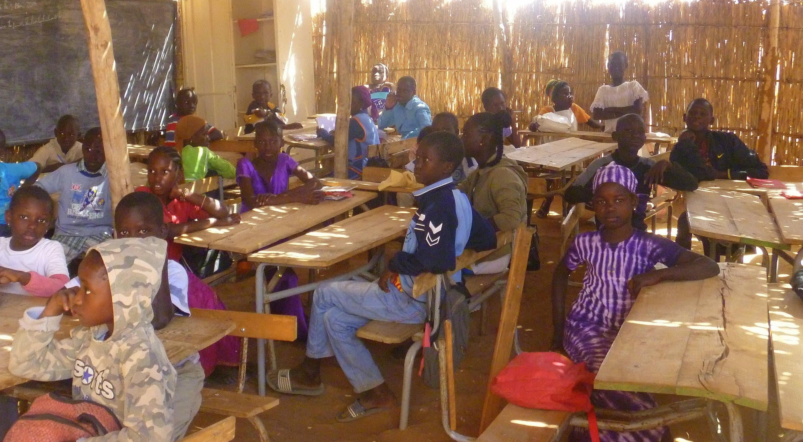 Children sitting in a makeshift classroom with reed walls and a dirt floor