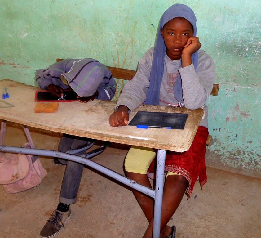 Two children sitting at a worn desk with slate boards, working in a run-down classroom